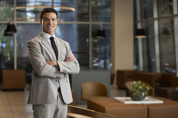 An young MBA graduate dressed in a suit stands smiling with arms crossed. 