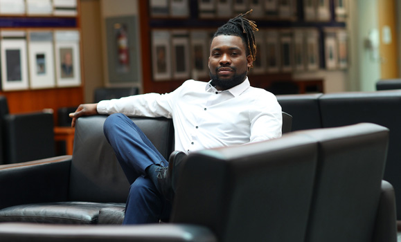 Mona Agada, a black man with a short beard and moustache, sits on a sofa in the lobby of the Rowe building. He is leaning back with one arm draped on the back of the sofa, and he has one foot resting on his knee in a relaxed position. He is wearing a white buttoned shirt and jeans. His medium-length locs are gathered into a high ponytail.