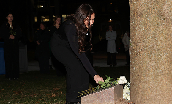 student wearing black places a white rose at the base of a tree