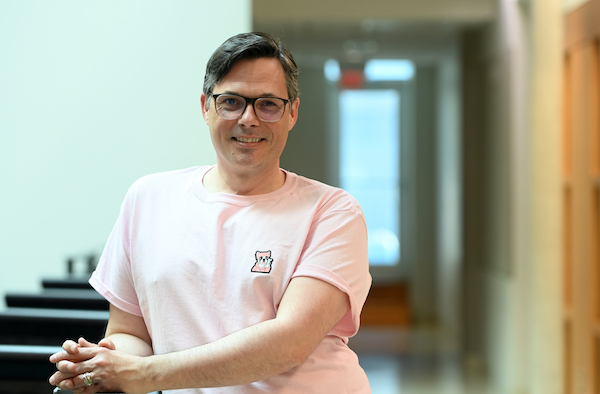 Jason Swinamer, a white man with glasses and short dark hair parted on one side, stands leaning against a railing with his hands clasped. He is wearing a pink t-shirt with a small cartoon dog in the top corner and smiling while looking into the camera. 