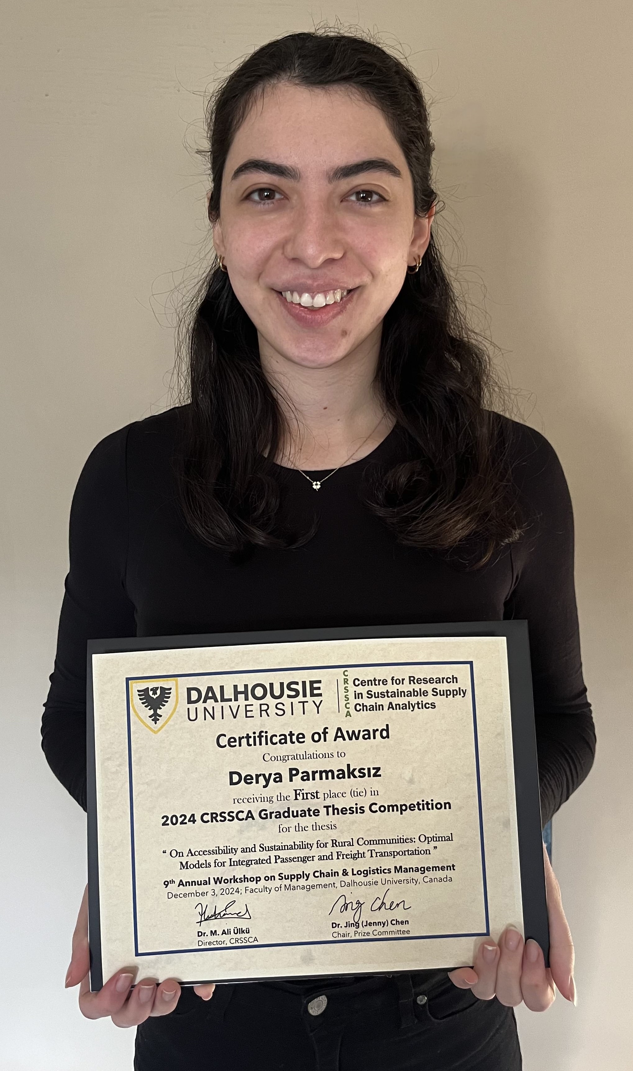 Derya stands holding a certificate, smiling at the camera. 