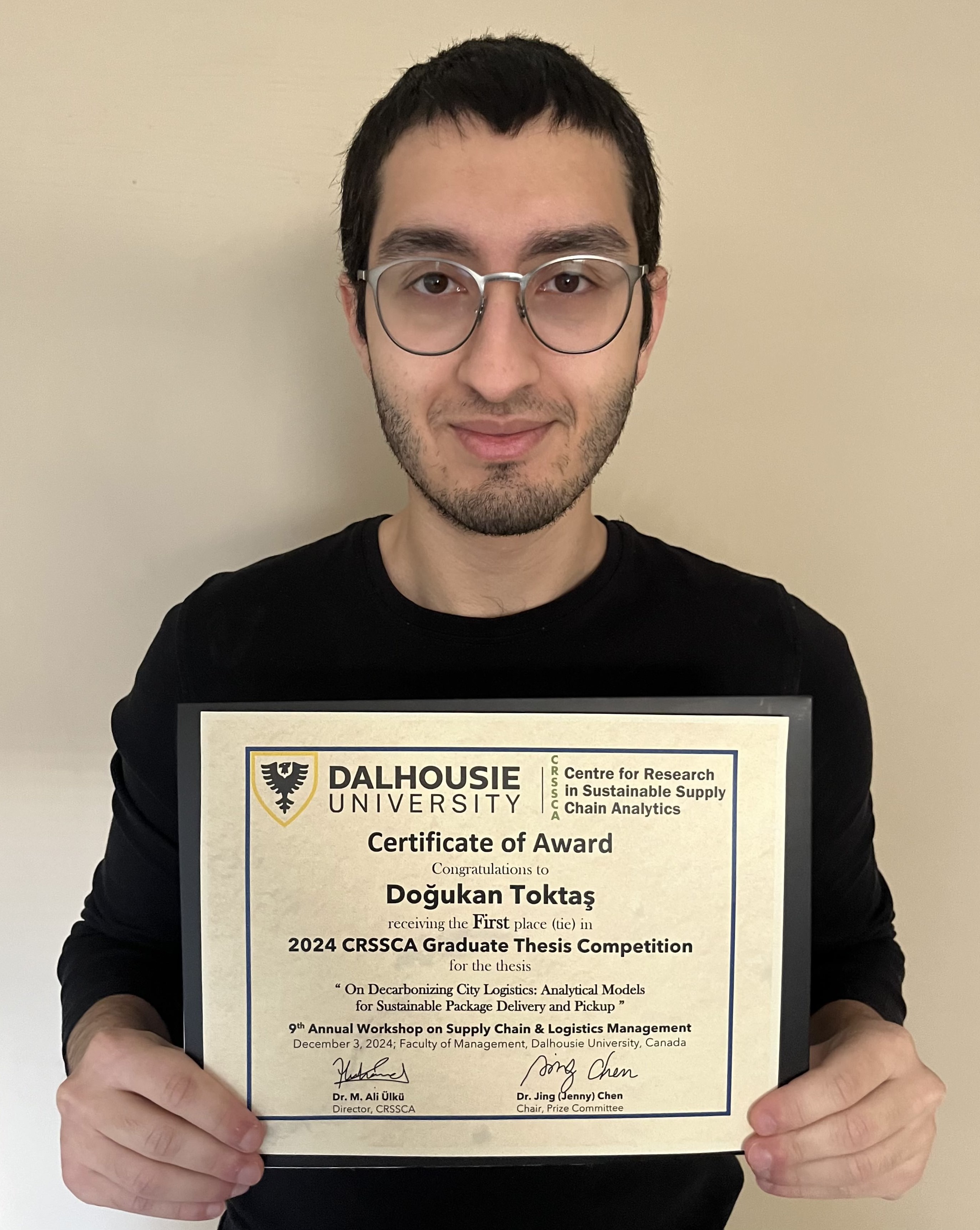 Doğukan stands holding a certificate, smiling at the camera. 