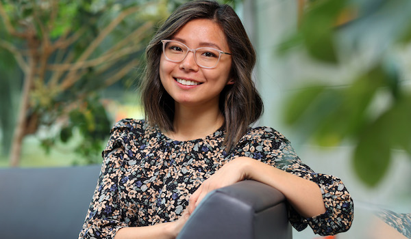 Byungmin Kang, a Korean woman with medium-length brown hair featuring light highlights, sits sideways on an armchair in the lobby of the Rowe building in front of a panel window with trees visible in the background. She is wearing a floral top and glasses, and smiling at the camera with one arm rested on the back of the armchair in front of her. 