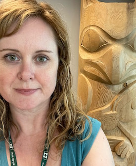 Katie Moran, a white woman with wavy blonde highlighted hair wearing a turquoise shirt, in front of an Indigenous wood carving. 