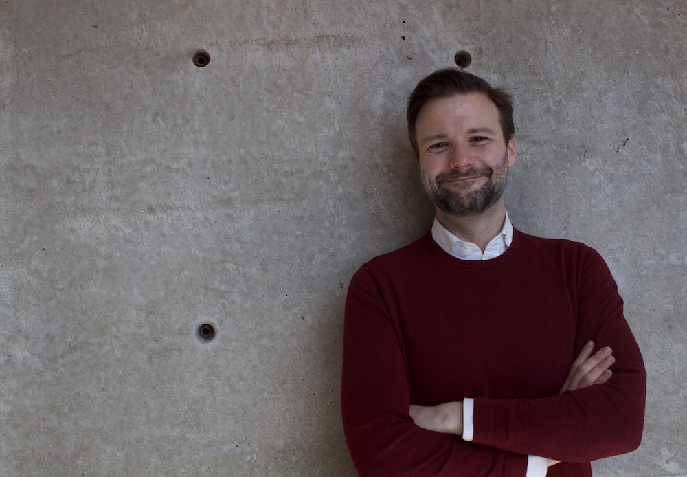 Andrew is leaning against a wall, arms crossed and smiling. He has light toned skin, short dark hair, and the beginnings of a beard and moustache.. 