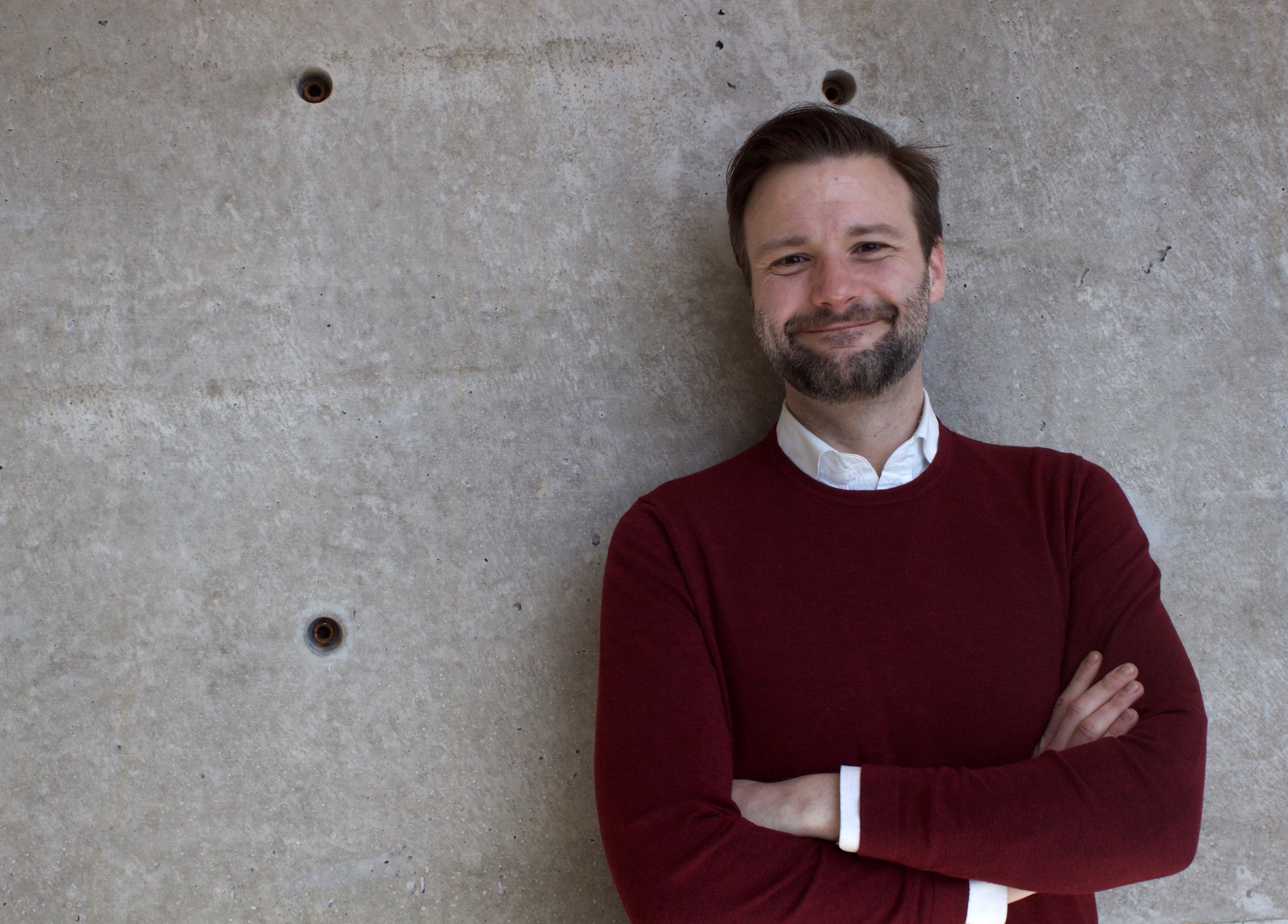 Andrew is leaning against a wall, arms crossed and smiling. He has light toned skin, short dark hair, and the beginnings of a beard and moustache.. 