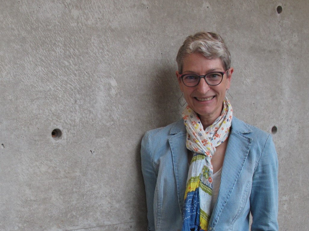 Georgia smiles at the camera. She has light-toned skin, short-cropped grey hair, and glasses, and she is wearing a blue jacket and colourful scarf.. 