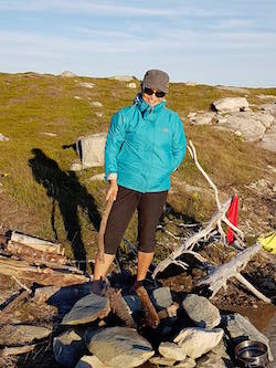 San stands on a hillside near a fire pit, dressed in a windbreaker, hat and sunglasses.