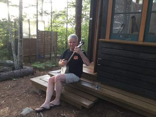 Steve strums a banjo while sitting on the steps of a cottage.