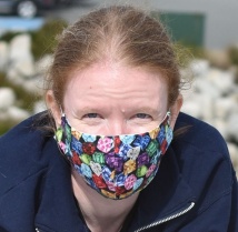 Lexie has a light skin tone and brown hair tied back in a ponytail. She is wearing a multi-coloured face mask and a dark blue shirt.