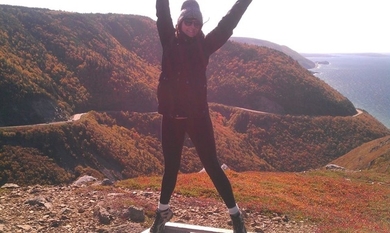 Laurel stands outdoors on a sunny hillside, her arms outstretched to either side of her, with the sea visible in the distance.