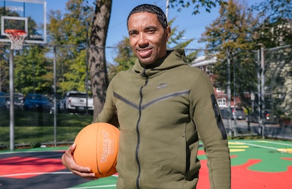 Rodney Small stands on an outdoor basketball court in a green sweatshirt, holding a basketball and smiling. 