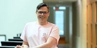 Jason Swinamer, a white man with glasses and short dark hair parted on one side, stands leaning against a railing with his hands clasped. He is wearing a pink t-shirt with a small cartoon dog in the top corner and smiling while looking into the camera.