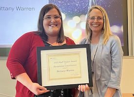 Brittany Warren holds her award certificate. She is standing next to Anna Cranston and they are both smiling at the camera.