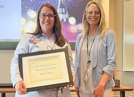 Kim Humes holds her award certificate. She is standing next to Anna Cranston and they are both smiling at the camera.