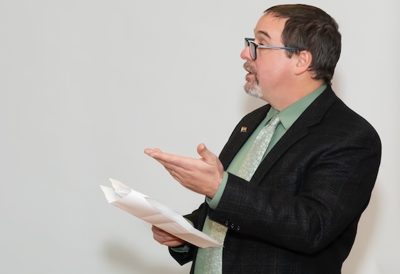Fred faces off-screen, gesturing as he speaks. He holds a copy of a speech.