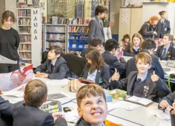 A group of students around a table with bookshelves in the background. One gives a thumbs up and one makes a face at the camera.