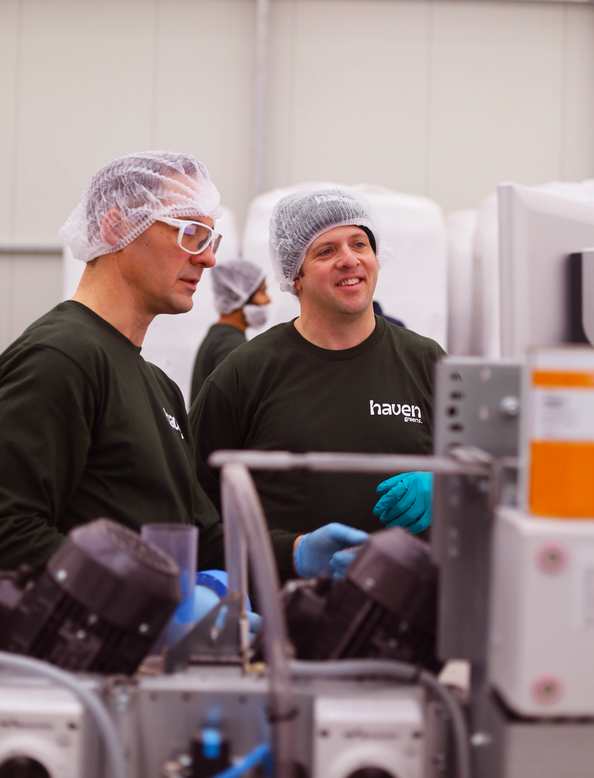 Jay and a coworker wearing blue globes and hairnets, operate machinery together.