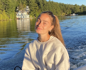 Francesca sits in a boat with water and trees in the background. She is wearing a white sweatshirt and her hair is pulled back in a long ponytail.