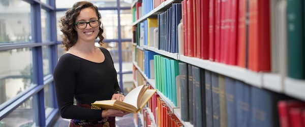 An MI student stands holding an open book next to a row of bookshelves in an academic library. 