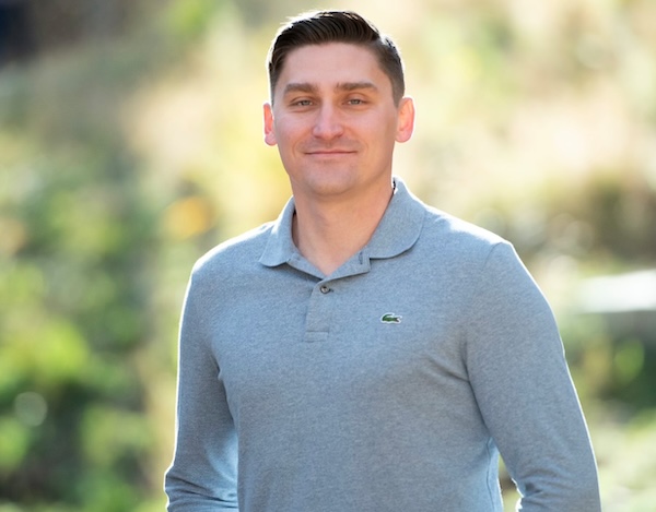 Stephen smiles at the camera, wearing a light blue collared shirt. He is outside with tress in the background. 