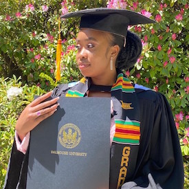 Kayler stands outside, surrounded by green leaves and pink flowers. She is wearing her graduation cap, robe and kente sash and holding her diploma.