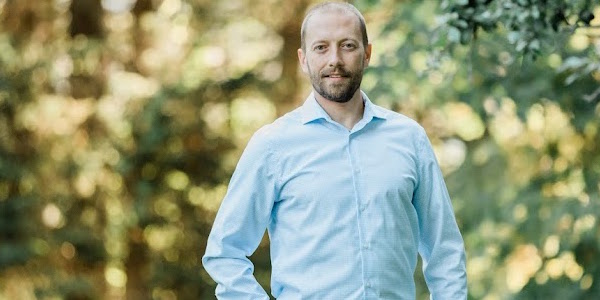 Patrick Law, a man with short brown hair, a moustache and beard, stands under a tree with one hand in his pocket. He is wearing a light blue button-up shirt and blue jeans and smiling while looking directly into the camera. 