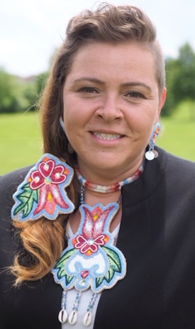 Christa Big Canoe, an Anishinabek woman, is shown outdoors, smiling at the camera, wearing beaded regalia and a black top. Her long hair is tied in a ponytail on one side. 