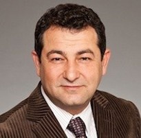 Photo of Anne Camozzi. Woman with long straight grey hair, glasses, wearing blue sweater and scarf with blue painting in background