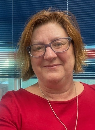 Photo of Anne Camozzi. Woman with long straight grey hair, glasses, wearing blue sweater and scarf with blue painting in background
