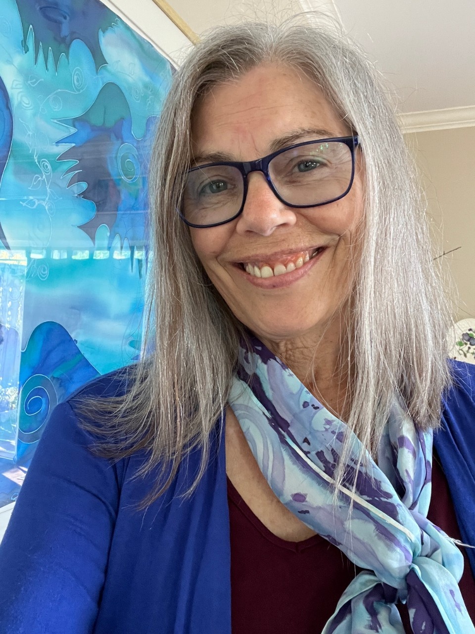 Photo of Anne Camozzi. Woman with long straight grey hair, glasses, wearing blue sweater and scarf with blue painting in background