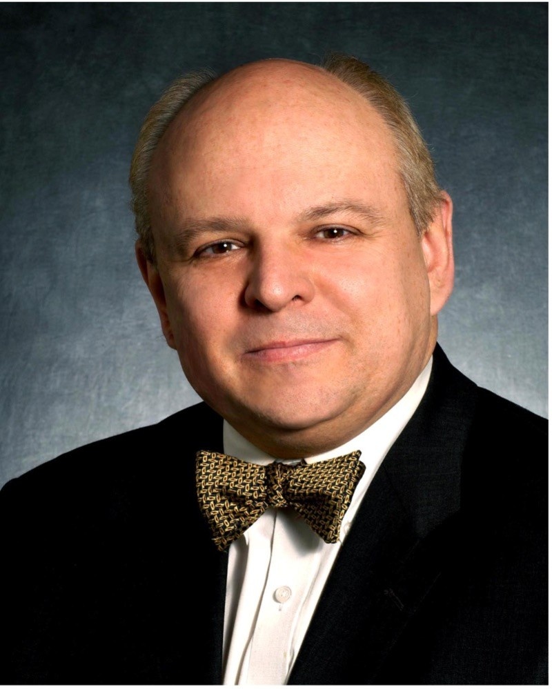 Photo of Paul Kovacs, professional studio photo with grey background, he is wearing white shirt, black jacket, and brown bow tie