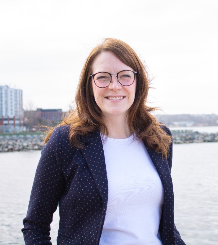 Photo of Mayor McDougall-Merrill. Long brown hair, white shirt, blue dotted jacket, she is pictured on waterfront