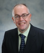 Photo of Kevin Quigley, he is smiling, wearing glasses, a green shirt and blue tie with a black suit