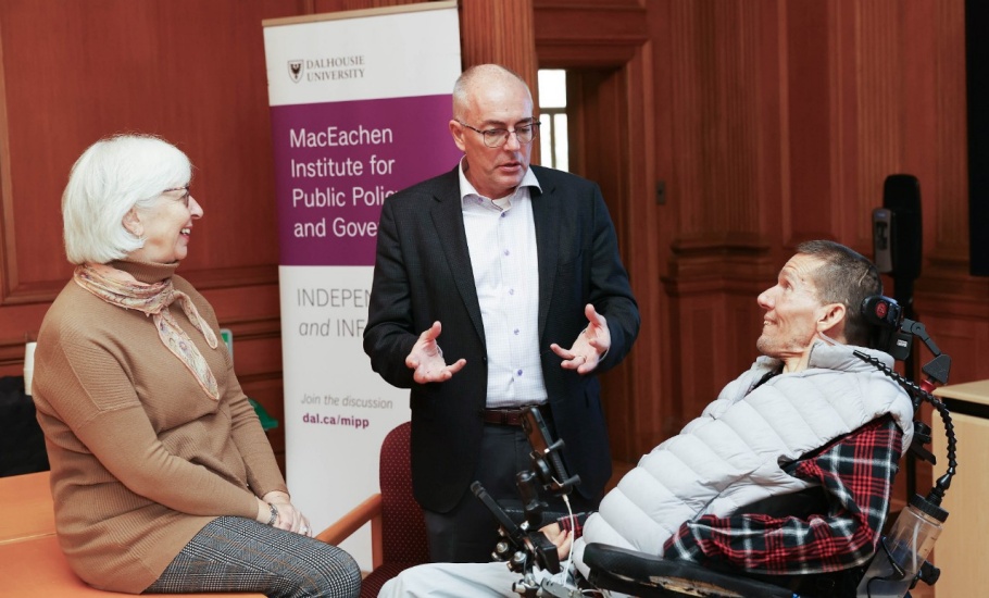 A man wearing glasses and a black suit stands between two people having a discussion. To his left is a woman sitting down. She has white shoulder-length hair and is wearing a brown sweater. To his right is a man sitting in a power wheelchair. He is wearing a red plaid shirt and grey vest.