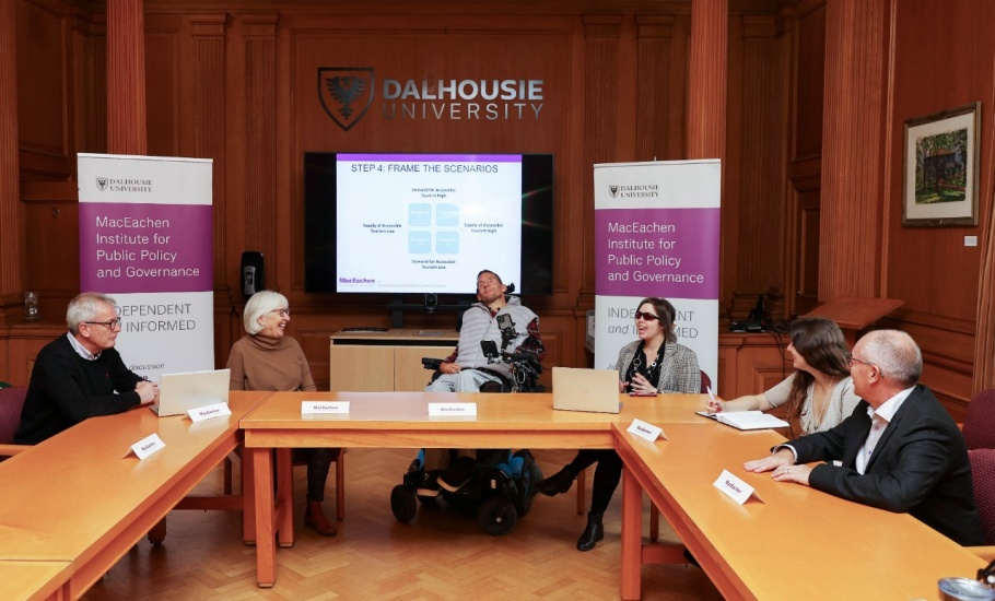 Six people are sitting around a table looking towards one person at the front of the room. They are each sitting behind a placard reading “MacEachen Institute”. At the front of the room is a projector screen that reads “Step 4: Frame the Scenarios” followed by a graph. Above the screen is a wall decal that reads “Dalhousie University”. The screen is flanked by two tall purple and white banners reading “MacEachen Institute for Public Policy and Governance”.