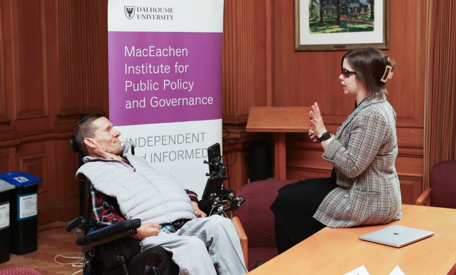 A woman and a man are sitting in front of a purple and white banner reading “MacEachen Institute for Public Policy and Governance”. The man is on the left, and is sitting in a power wheelchair. He is wearing a red plaid shirt and grey vest. The woman is on the right, and is wearing a long grey plaid blazer and black pants. She is speaking and gesturing with her hands. She is wearing dark glasses.