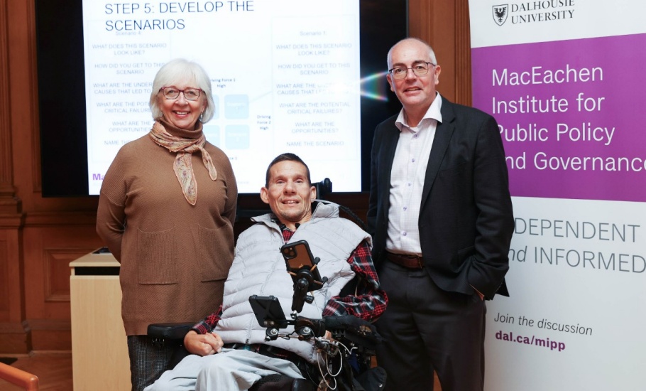 Three people are in front of a projector screen and a purple and white banner. The projector screen reads “Step 5: Develop the Scenarios” and the banner reads “MacEachen Institute for Public Policy and Governance”. Standing on the left is a woman with white hair and a brown sweater. In the centre, a man is sitting in a power wheelchair. He is wearing a red plaid shirt and grey vest. Standing on the right is a man with glasses. He is wearing a black suit. All three are smiling.