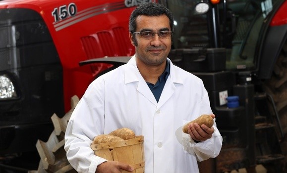 Dr. Ahmad Al-Mallahi is photographed wearing a lab coat and protective eyewear standing in front of a tractor holding a basket of potatoes.