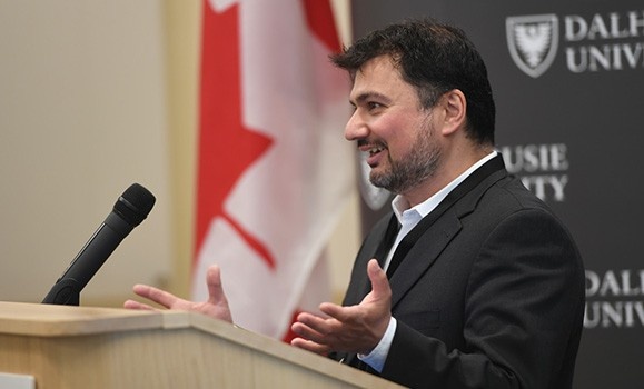 A photograph of Jordan Kyriakidis standing at a podium by a Canadian flag
