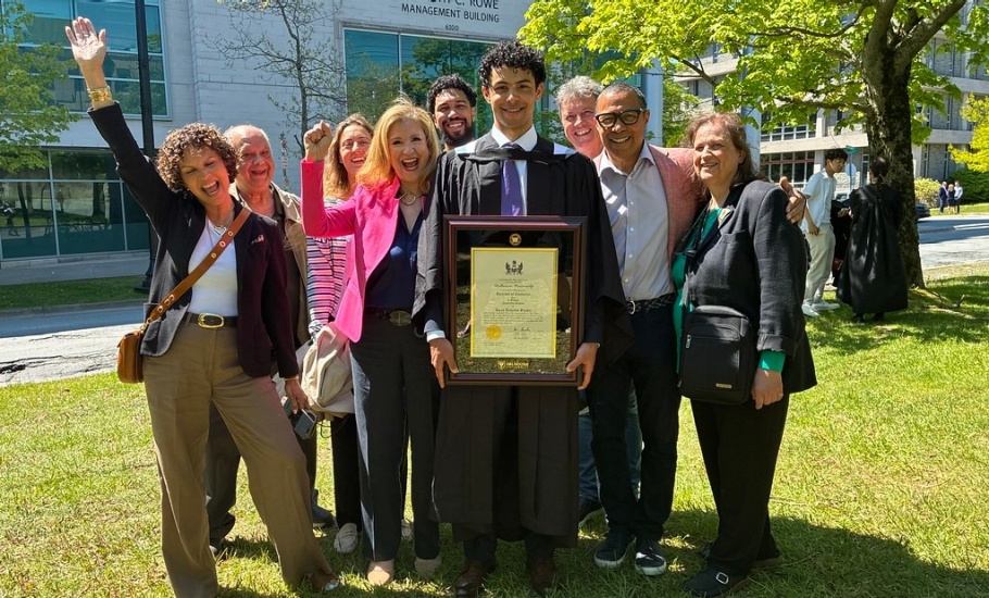 A graduate in gown stands outside in front of the Rowe building with family behind him cheering.