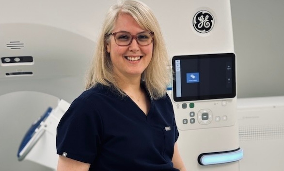 Lesley poses in front of a CT machine wearing navy blue scrubs and smiling.