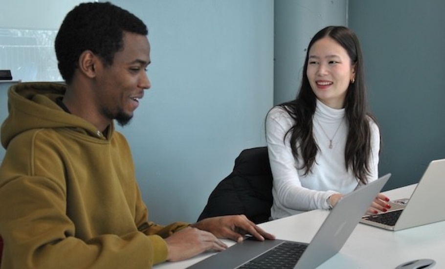 Two students sit at a table with laptops, engaged in conversation with each other.