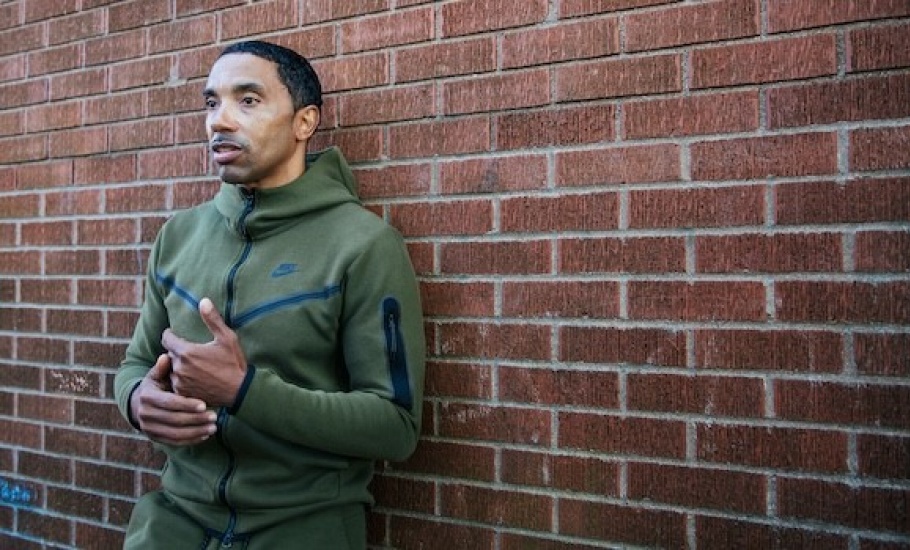 Rodney leans against a brick wall wearing a green sweatshirt. He is gesturing as though having a conversation with someone offscreen.