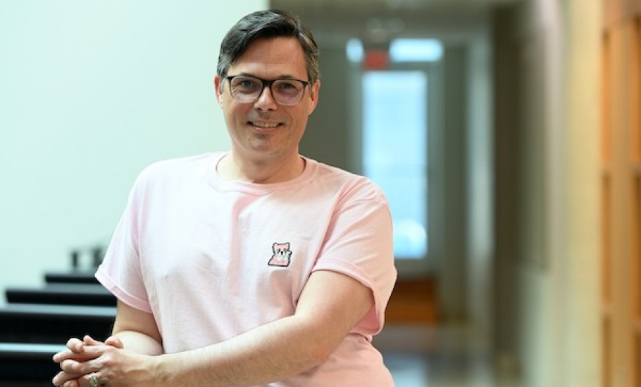 Jason Swinamer, a white man with glasses and short dark hair parted on one side, stands leaning against a railing with his hands clasped. He is wearing a pink t-shirt with a small cartoon dog in the top corner and smiling while looking into the camera.