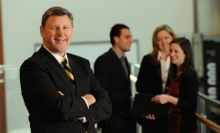 A man faces the camera,smiling. In the background, 3 others' are having a discussion.