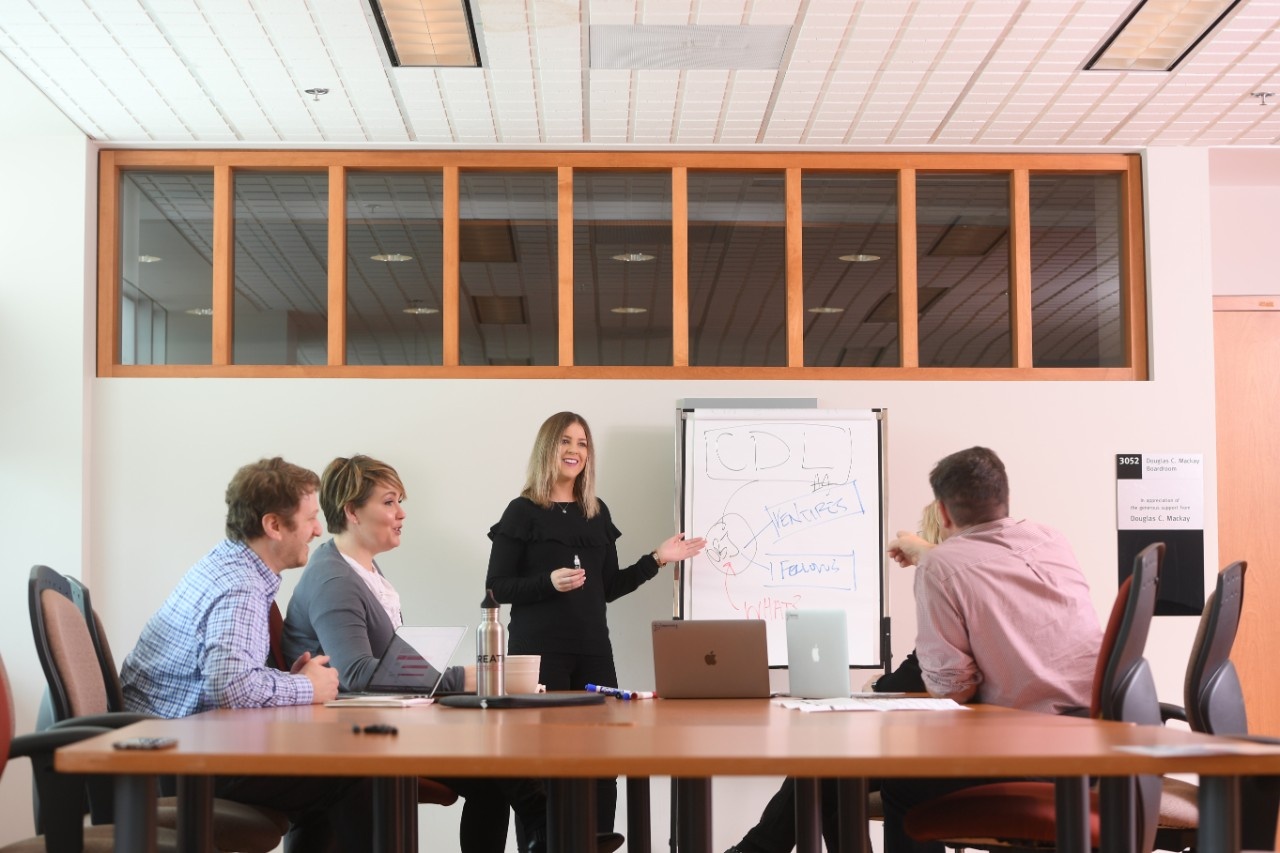 Four professionals sit around large board room table while woman presents at easel.