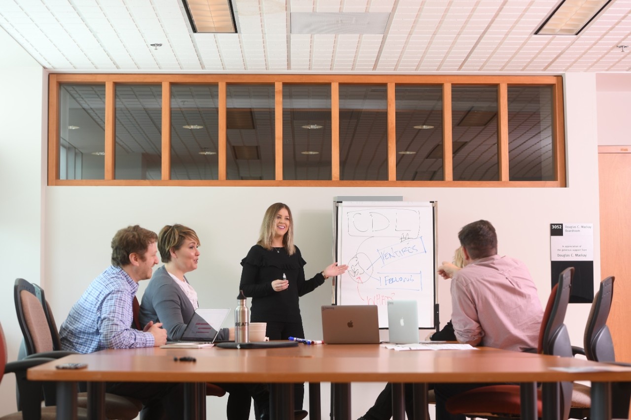 Four professionals sit around large board room table while woman presents at easel. Four professionals sit around large board room table while woman presents at easel.