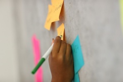 A hand is shown writing on a sticky note, which is posted to a wall during a learning exercise.