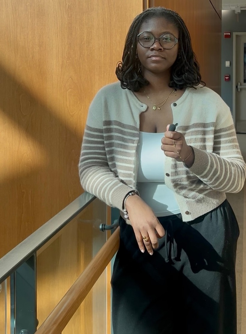 Dumebi leans on a balconey railing, holding a microphone and wearing a striped cardigan.